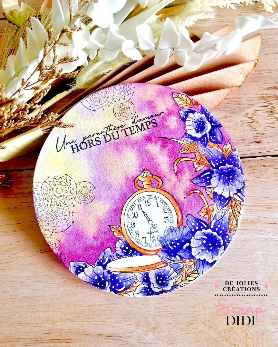 Une carte ronde en aquarelle florale avec tampon horloge et fond pastel. Cette création est une aquarelle ronde unique et poétique, parfaite pour les amoureux de scrapbooking et de carterie créative. Sur un fond délicat mêlant roses, jaunes et violets à l’aquarelle, une composition florale vient encadrer un tampon horloge vintage, évoquant une parenthèse hors du temps.  Les fleurs bleues peintes à l’aquarelle, rehaussées de touches de blanc au stylo acrylique, apportent relief et douceur. La citation “Une parenthèse d’amour hors du temps” renforce l’aspect émotionnel et intemporel de la création.  Cette carte a été réalisée en utilisant la technique du cache pour superposer les éléments sans surcharge visuelle : un procédé essentiel en tamponnage créatif pour créer des effets de profondeur.